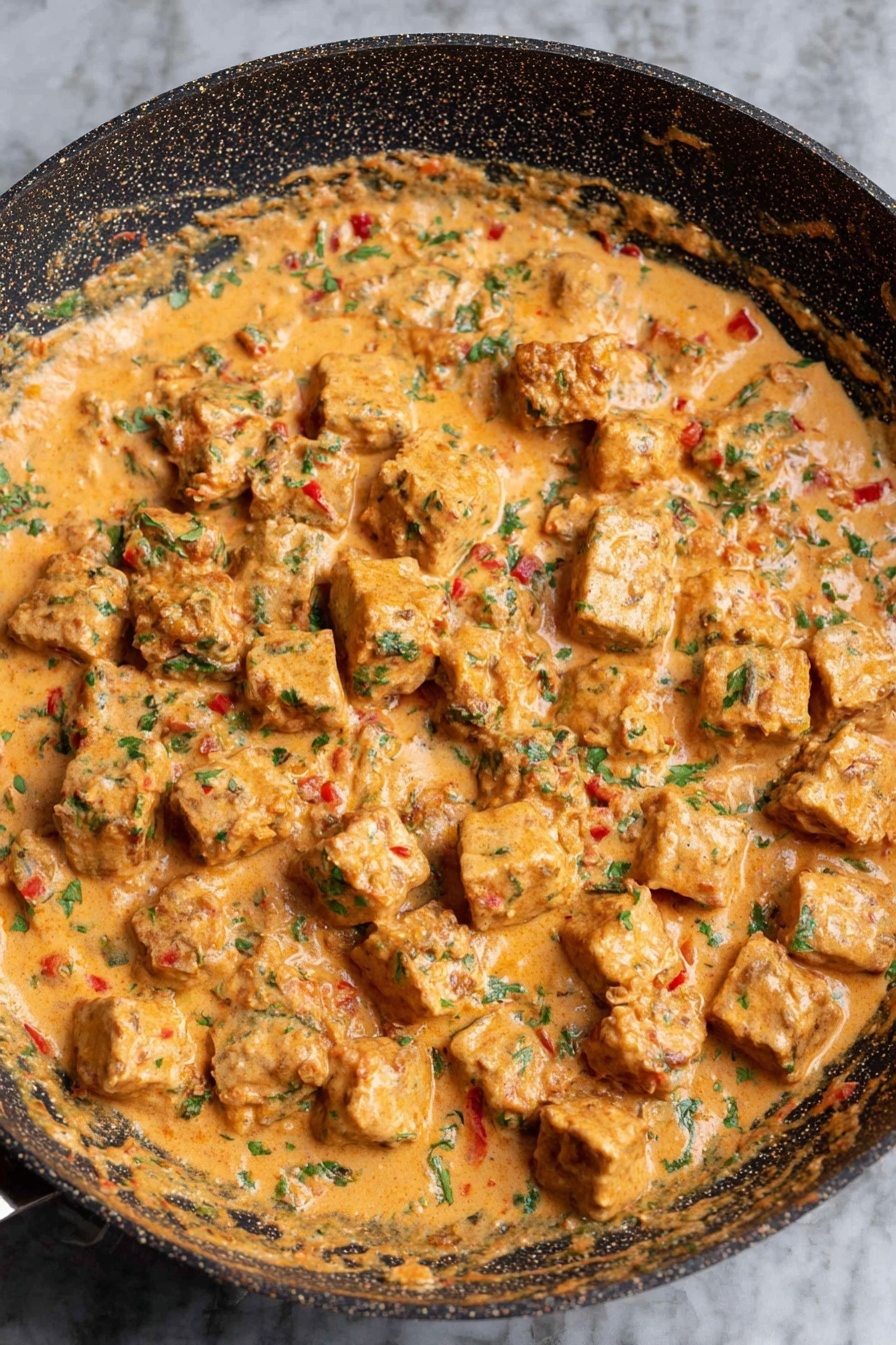 Sun-Dried Tomato Cashew Cream - The image shows a pan filled with a creamy sauce that has a light orange color mixed with green herbs and small red bits. In the sauce, there are many evenly sized, light brown cubes that are partly covered by the sauce. The pan is round with a dark, patterned surface, and it sits on a white marbled background. The texture of the sauce looks thick and smooth with visible small pieces blending in photo taken with an iphone --ar 2:3 --v 7