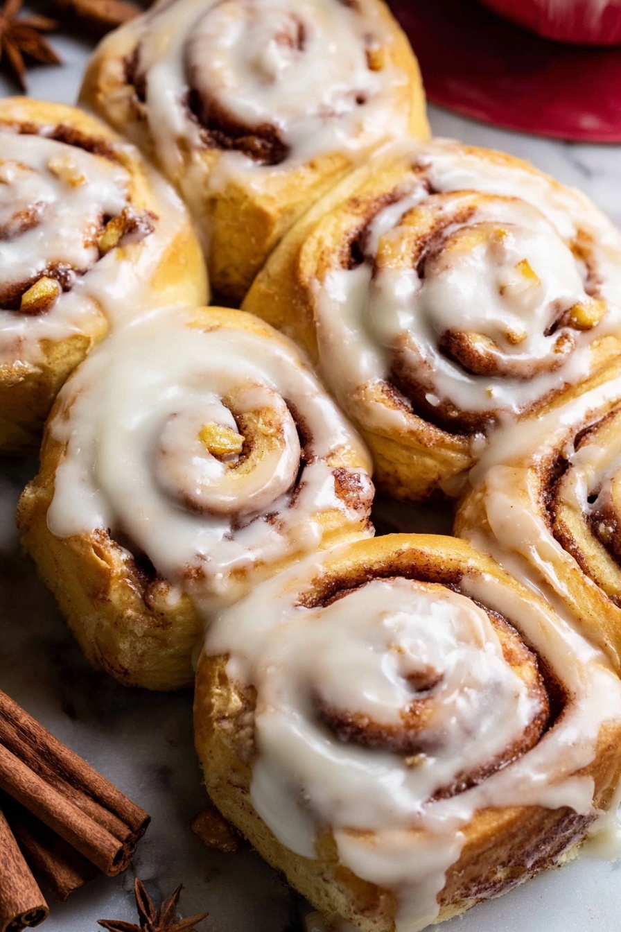 The image shows six cinnamon rolls arranged closely together on a white marbled surface. Each roll has several spiral layers, with golden-brown dough and visible cinnamon filling, some pieces of chopped nuts or apple bits are inside the spirals. A thick layer of creamy white icing covers the top of every roll, dripping slightly down the sides and filling some of the inner swirls. The background includes blurred cinnamon sticks and a bit of a red object in the upper right corner. The rolls look soft, warm, and freshly baked, with a slightly shiny icing texture. photo taken with an iphone --ar 2:3 --v 7