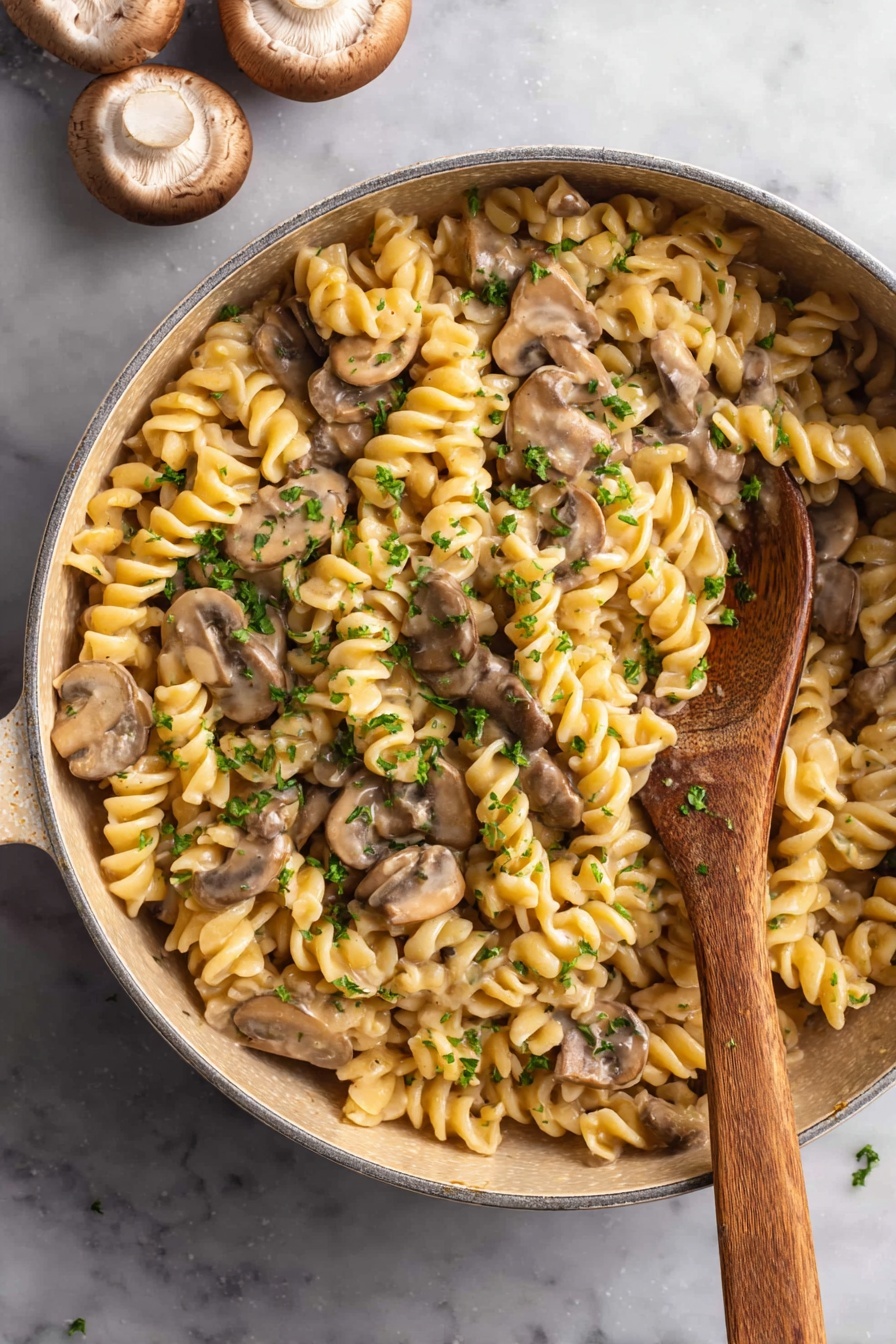 Vegan Mushroom Stroganoff - The image shows a round light beige pot filled with creamy rotini pasta and sliced mushrooms mixed together. The twisted pasta is pale yellow while the mushrooms are medium brown with lighter edges. Small pieces of green parsley are sprinkled over the pasta. A wooden spoon is inside the pot, partially covered with the pasta dish. The pot is placed on a white marbled surface with two whole mushrooms seen in the top left corner. Photo taken with an iphone --ar 2:3 --v 7