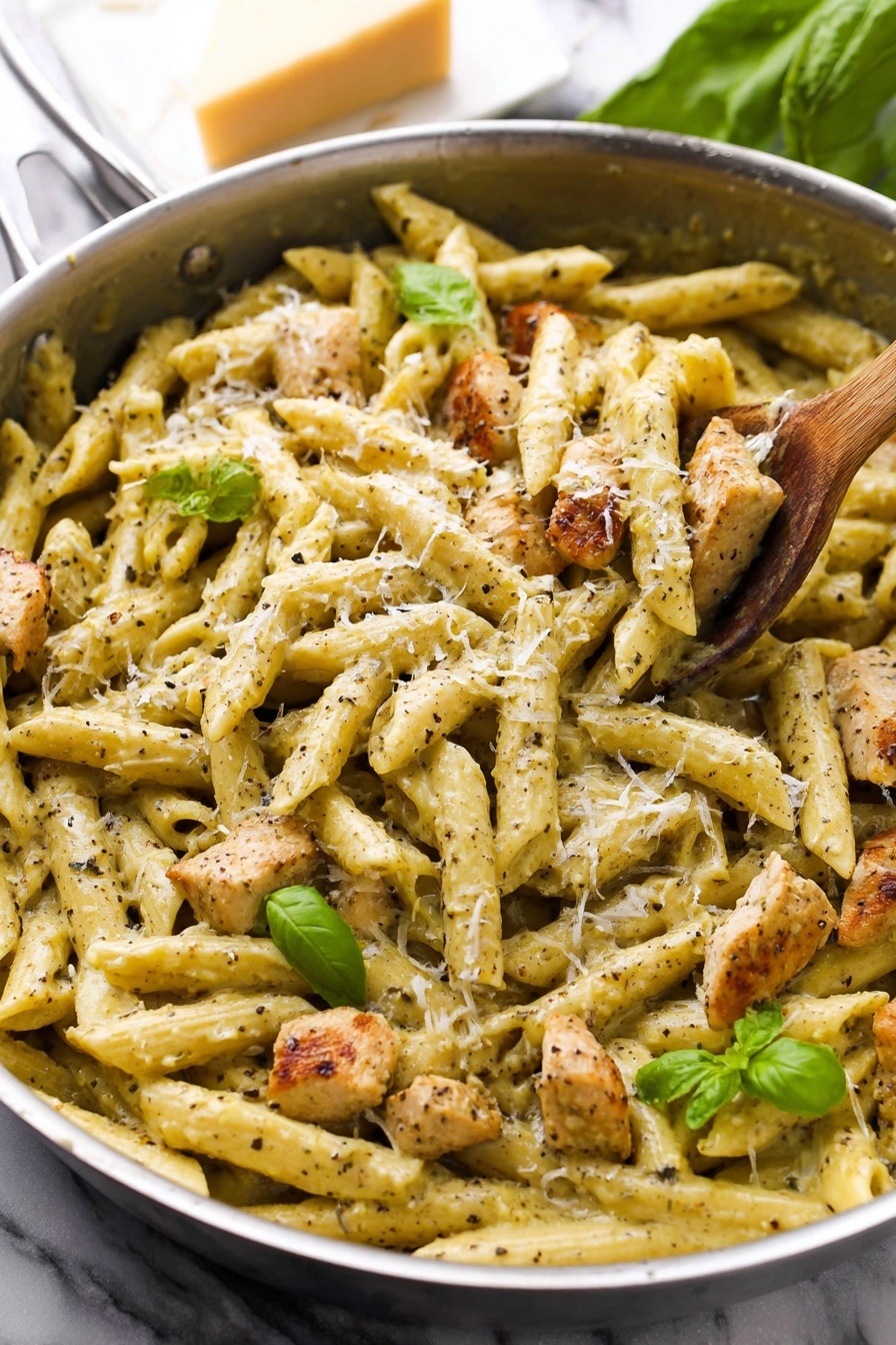 Creamy Pesto Chicken - A close-up view of a stainless steel pan filled with creamy penne pasta mixed with browned pieces of seasoned chicken. The pasta is coated in a pale yellow sauce with visible black pepper and herbs throughout. Small bright green basil leaves are scattered on top, along with finely grated white cheese sprinkled over the dish. A wooden spoon rests inside the pan, partially lifting some pasta, showing the thick creamy texture. The pan is placed on a white marbled surface with a block of cheese and some green leaves blurred in the background. Photo taken with an iphone --ar 2:3 --v 7