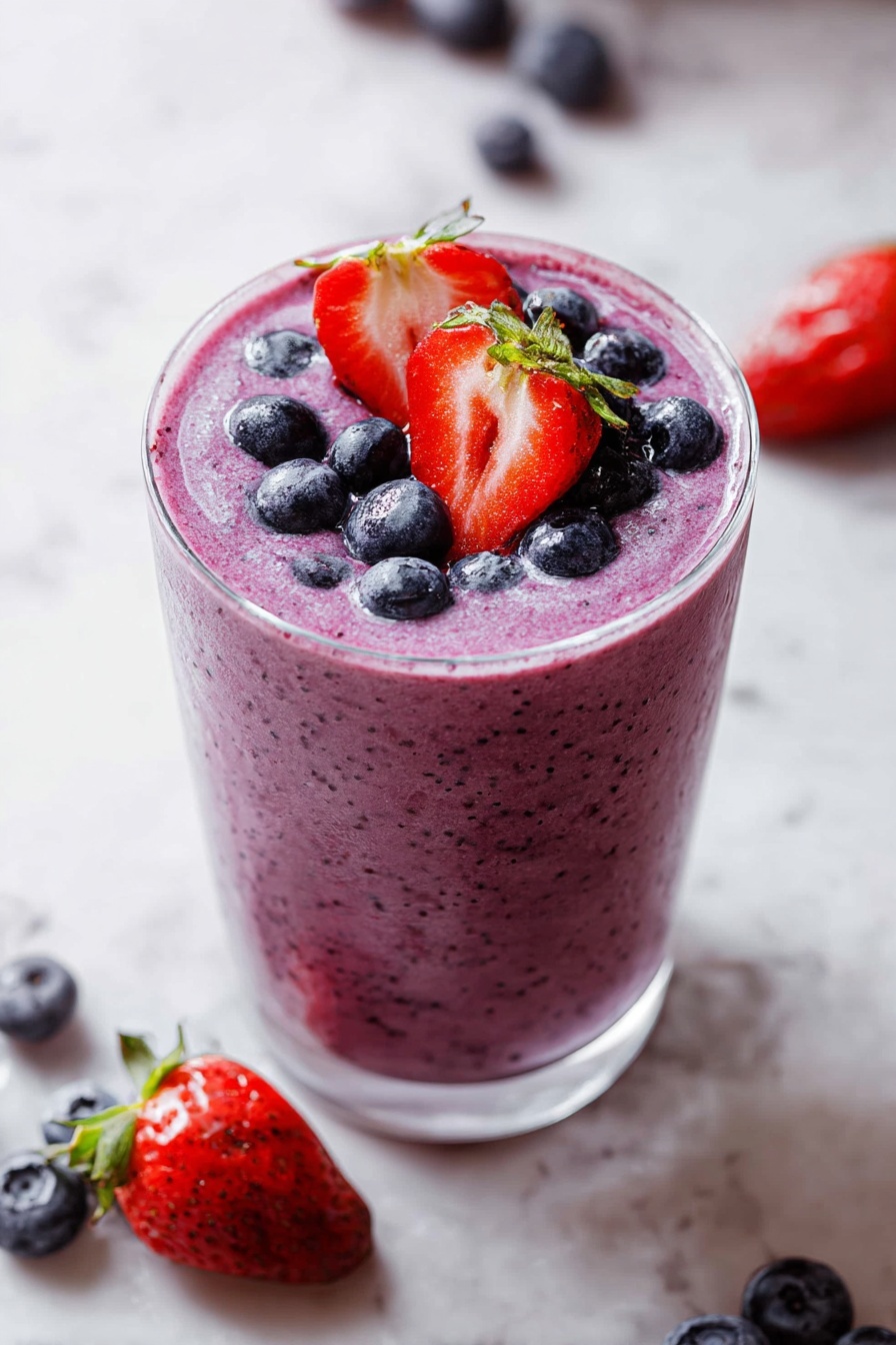 A tall clear glass filled with a thick, purple smoothie with tiny dark specks inside. The top layer is decorated with whole, dark blue blueberries scattered around and two halves of bright red strawberries with green tops standing up in the middle. The glass is on a white marbled surface with a few blueberries and a strawberry placed loosely around it. photo taken with an iphone --ar 2:3 --v 7