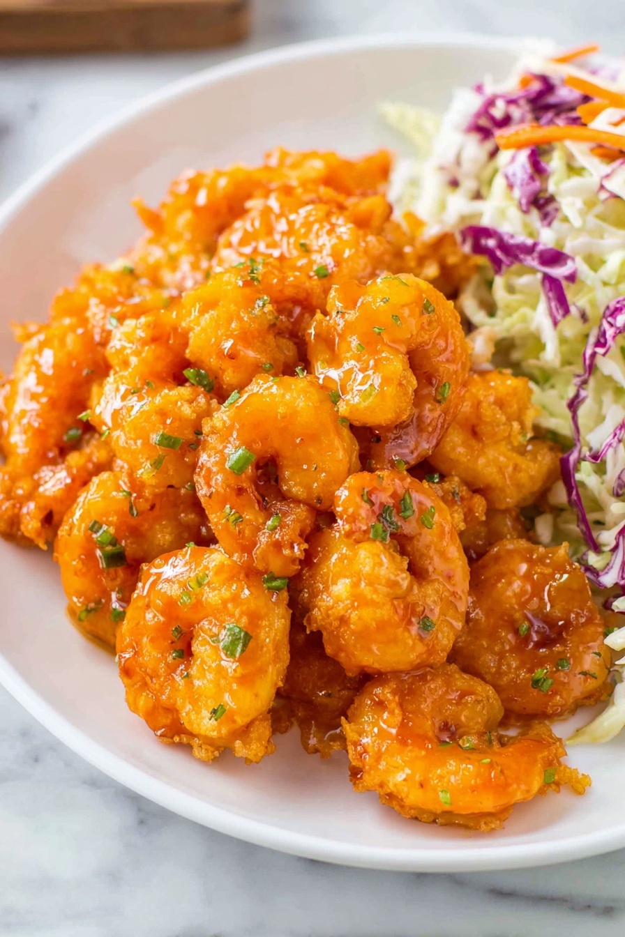 A white plate filled mostly with bright orange fried shrimp coated in a shiny sauce, slightly piled in the center and sprinkled with small bits of green herbs. In the background at the top right edge of the plate, there is a small serving of coleslaw made of shredded cabbage, some purple and white pieces mixed with a few thin slices of carrots. The plate sits on a white marbled surface. photo taken with an iphone --ar 2:3 --v 7