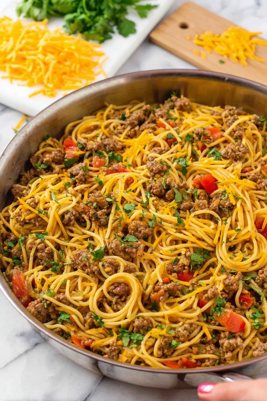 A large silver pan is filled with a mix of long, thin yellow spaghetti noodles and browned ground meat pieces. Scattered throughout are small, bright red tomato chunks and melted orange-yellow cheese strands that lightly cover the dish. Fresh green herb leaves are spread on top, adding a touch of color. In the background, there is a white marbled surface with a white cutting board holding a pile of shredded yellow cheese and some fresh green herbs. A woman's hand is reaching toward the pan from the right side. Photo taken with an iphone --ar 2:3 --v 7