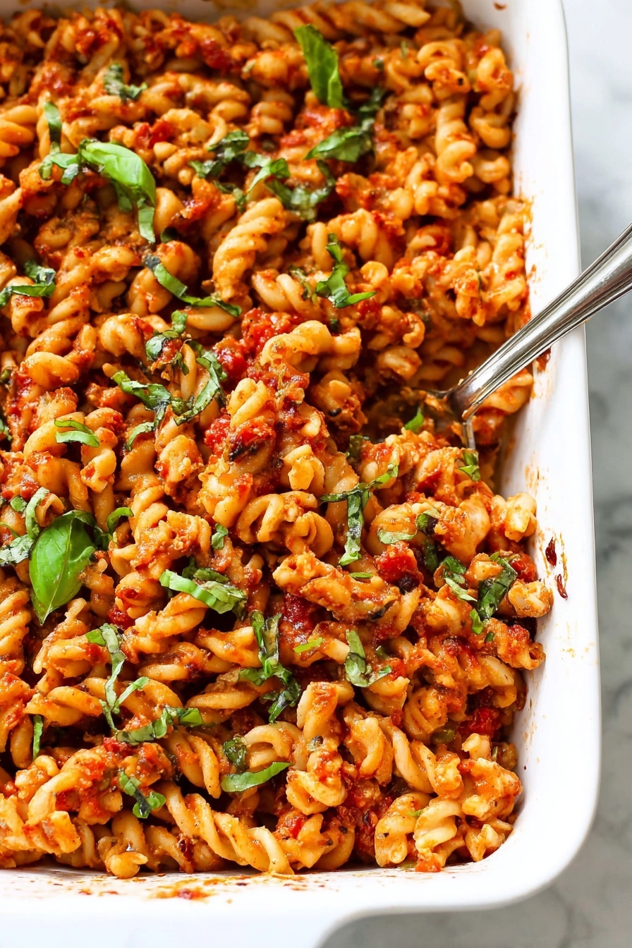 A white rectangular baking dish filled with a baked pasta dish made of twisted pasta shapes mixed with a red sauce that has visible pieces of tomato and herbs. The pasta is coated evenly with the sauce and sprinkled with chopped green basil leaves scattered on top, adding a fresh green color contrast. A silver spoon is placed inside the dish on the right side, partially submerged in the pasta. The surface underneath the dish is a white marbled texture. photo taken with an iphone --ar 2:3 --v 7