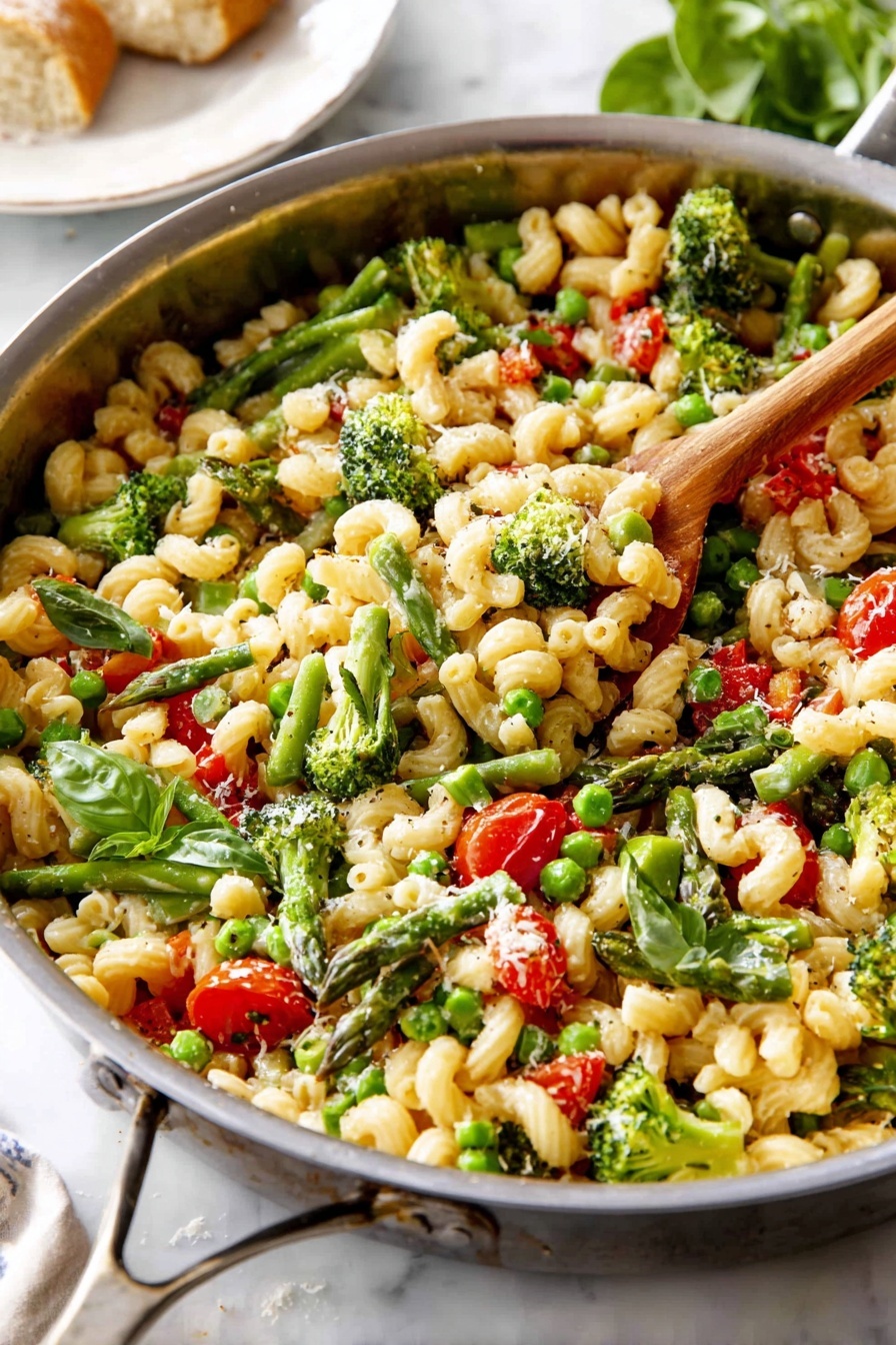 A large metal pan is filled with curly pasta mixed with bright green broccoli florets, peas, and asparagus cut into pieces. There are also red cherry tomato halves and small cubes of red bell pepper scattered throughout. Fresh green basil leaves are sprinkled on top, along with a light dusting of grated cheese and black pepper. A wooden spoon rests inside the pan, slightly lifting some pasta. The pan sits on a white marbled surface with a blurred white plate and bread in the background. photo taken with an iphone --ar 2:3 --v 7