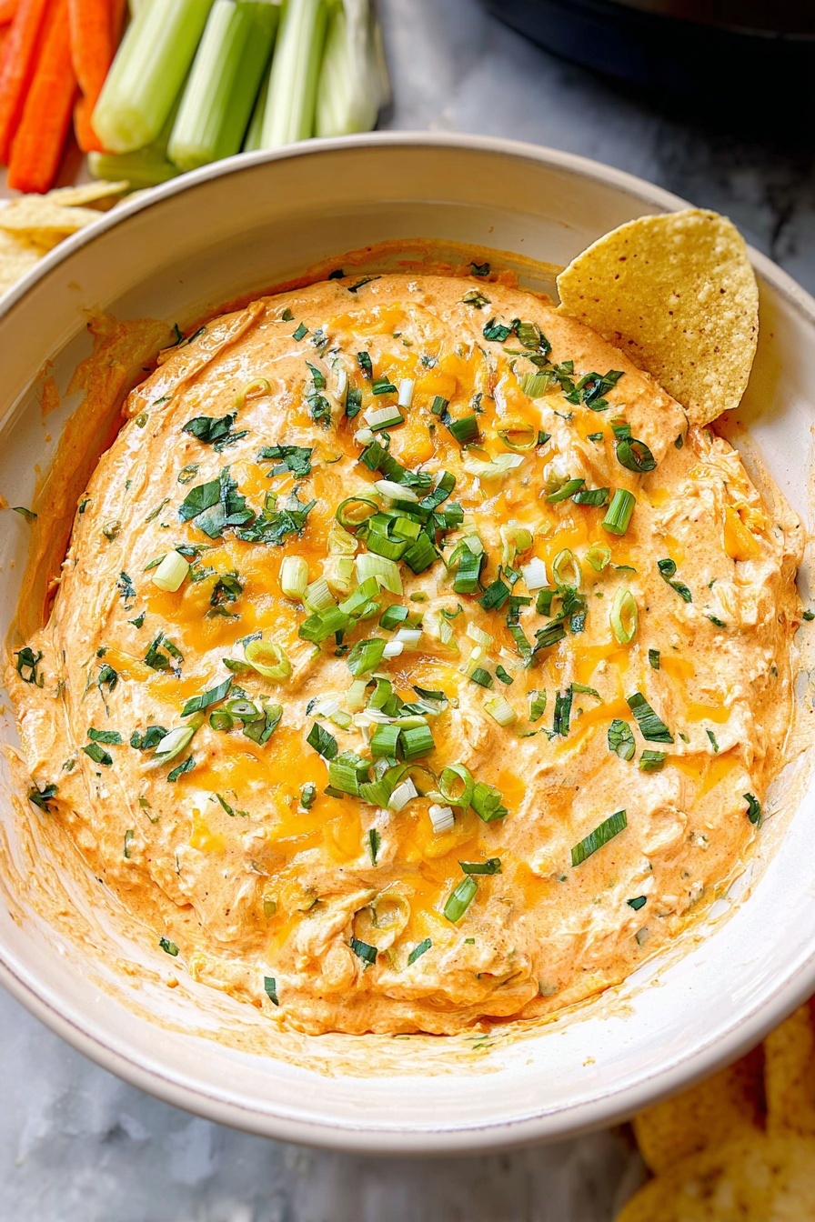 The image shows a thick, creamy dip inside a black slow cooker. It has a rich orange color with melted white and yellow cheese on top. There are small green pieces of chopped herbs and scallions sprinkled all over the dip. Two triangular light yellow chips are partially dipped into the right side of the dip. In the background, there are blurred bowls with orange carrots, green celery sticks, and more light yellow chips on a white marbled surface. photo taken with an iphone --ar 2:3 --v 7