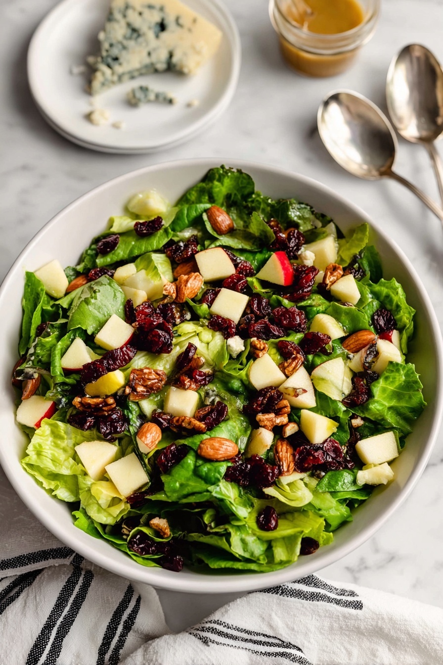 A white bowl filled with a fresh green salad made of large leafy greens as the bottom layer, topped with small cubes of pale yellow and light red apple pieces spread evenly across the salad. Scattered on top are brown roasted almonds and dark red dried cranberries, with small crumbles of blue cheese adding white with blue veins spots. In the background, a white plate with a wedge of blue cheese and two silver spoons can be seen on a white marbled surface along with a small jar of dressing. A white cloth with black stripes is partially visible at the bottom edge. Photo taken with an iphone --ar 2:3 --v 7
