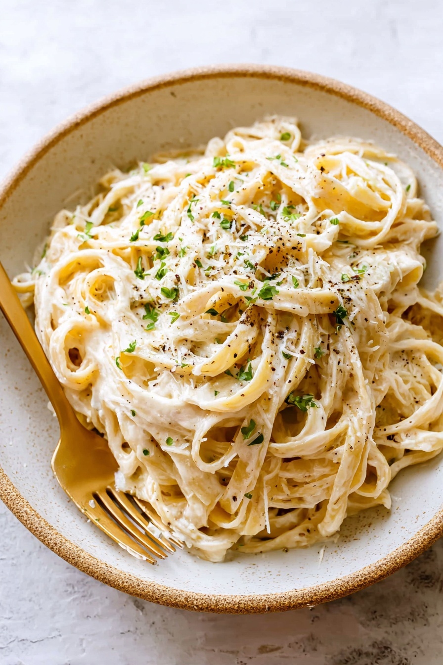 A large round white bowl holds a heap of creamy fettuccine pasta tossed in a smooth white sauce. The pasta strands are thick, coated evenly with the sauce, and topped with small pieces of green herbs and a sprinkling of black pepper that adds contrast and texture. A gold fork is inserted on the left side of the bowl, touching the pasta. The bowl sits on a white marbled surface that adds a soft, clean background to the dish. photo taken with an iphone --ar 2:3 --v 7