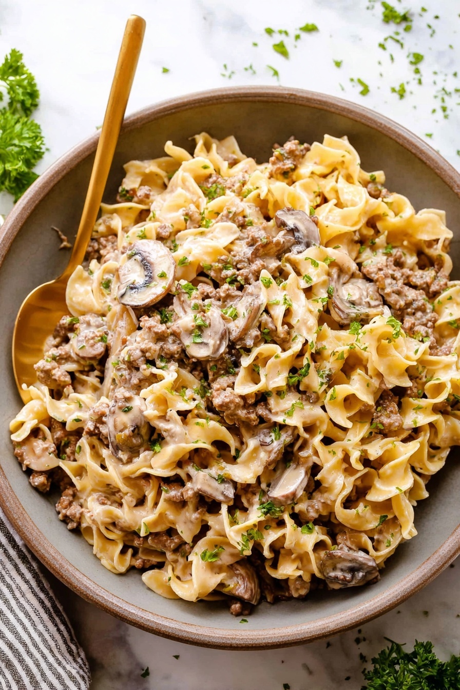 A close-up view of a bowl filled with creamy pasta made of wide, flat noodles layered with cooked ground meat and slices of browned mushrooms. The sauce is light-colored and coats all the pasta and meat evenly, with small green herb pieces sprinkled on top. The bowl is white and round, held by a woman's hand at the bottom, and a gold spoon is inserted into the side of the pasta. The background shows a white marbled surface with a striped cloth nearby. photo taken with an iphone --ar 2:3 --v 7
