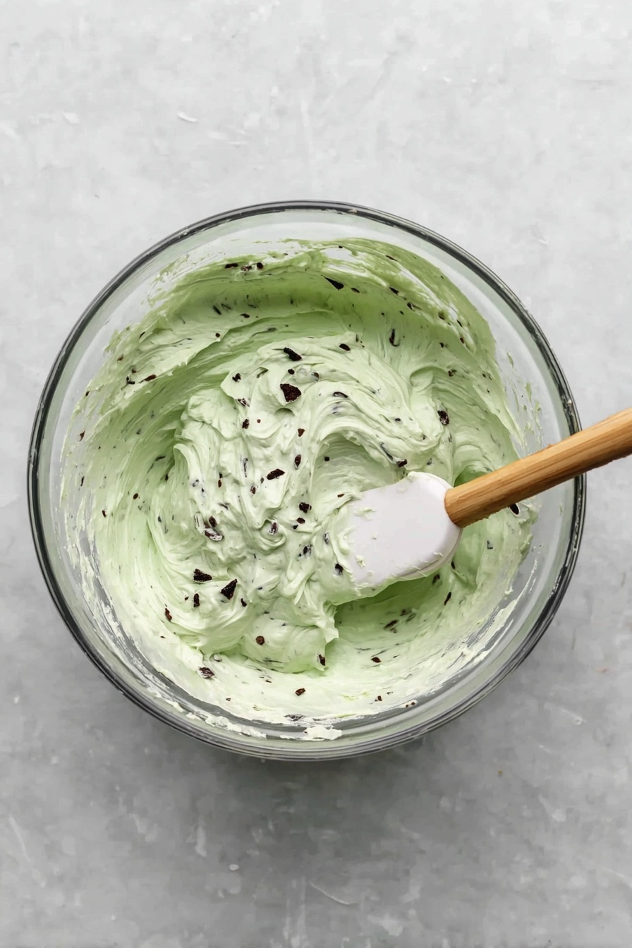 A slice of pie sits on a white plate with scalloped edges, placed on a white marbled surface. The pie has three visible layers: a dark chocolate crumb crust at the bottom, a thick middle layer of dark chocolate pudding, and a thick top layer of pale green mint cream mixed with small dark chocolate chunks. On top of the pie slice is a large swirl of white whipped cream, decorated with a dark chocolate curl and a fresh green mint leaf. Some crumbs are scattered on the plate near the slice. In the background, more pie is visible in a glass pie dish. photo taken with an iphone --ar 2:3 --v 7