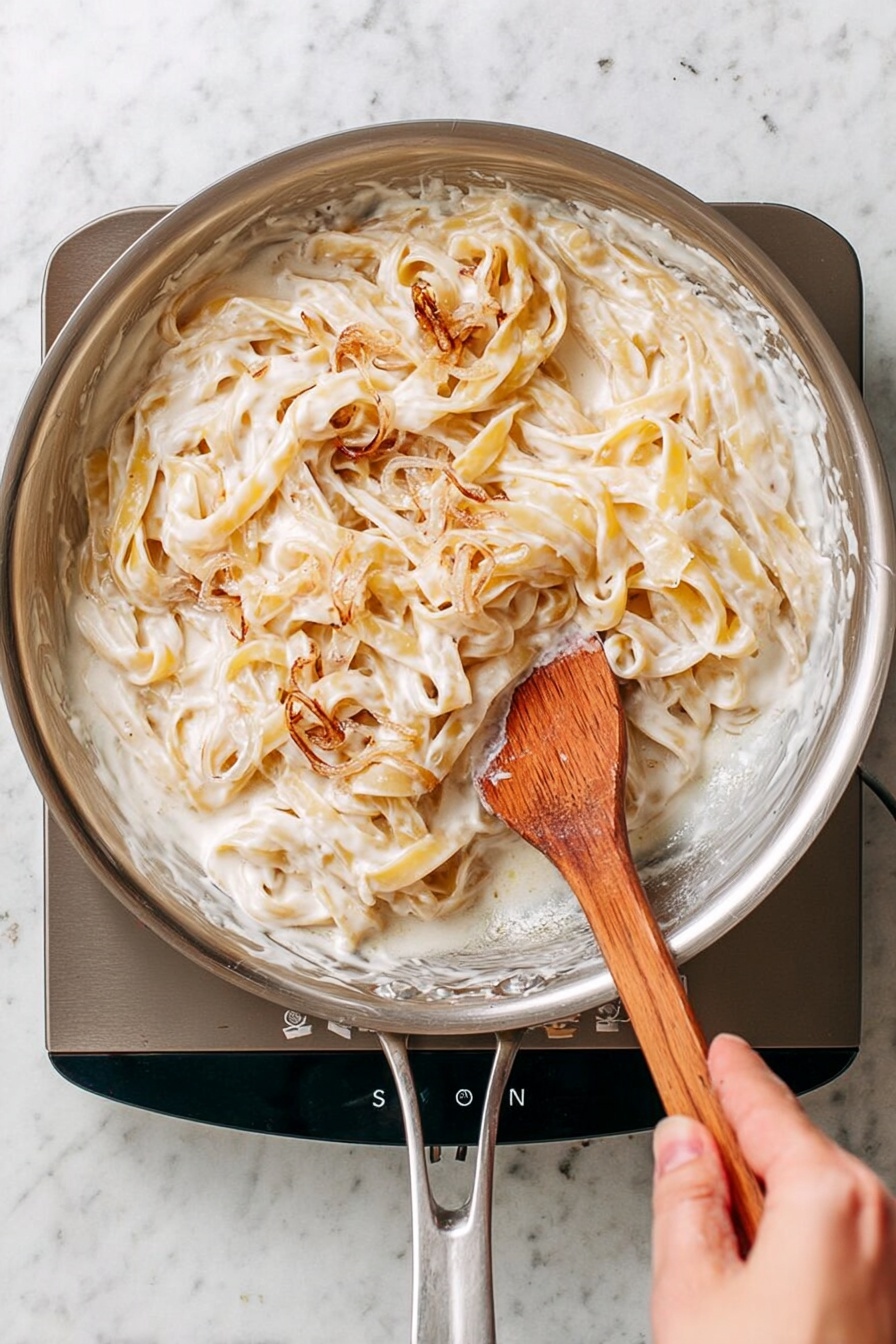A stainless steel pan filled with creamy white sauce coating light yellow fettuccine pasta, with some sauce slightly browned in spots. A woman's hand holds the pan handle at the bottom center, while the other woman's hand stirs the pasta with a wooden spatula on the right side, which has white sauce on its flat side. The pan sits on a sleek gray stove set on a white marbled surface. photo taken with an iphone --ar 2:3 --v 7
