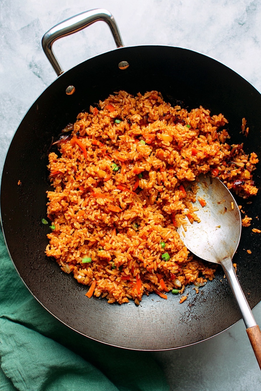 The image shows a black wok filled with a single layer of orange-colored fried rice mixed with small pieces of vegetables like onions and carrots, giving it a textured and slightly glossy look. The wok has a simple metal handle and a large metal spoon with some rice on it rests on the right side inside the wok. The background is a white marbled surface with a green cloth placed under the wok on the bottom left side. photo taken with an iphone --ar 2:3 --v 7