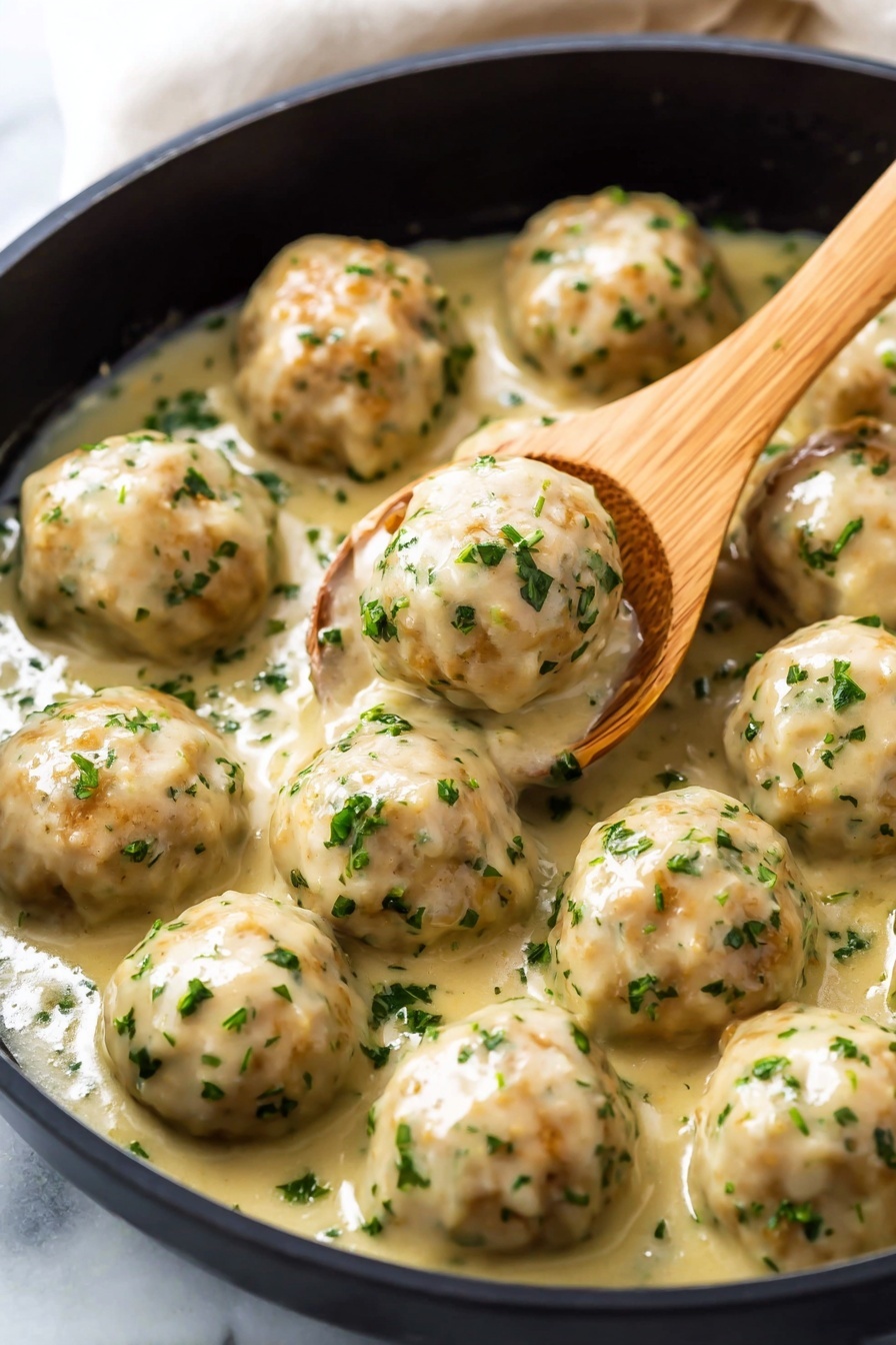 The image shows a close-up of a black skillet filled with about twelve round meatballs covered in a light cream sauce with small bits of green herbs visible on each meatball and in the sauce. The meatballs are smooth and round, coated evenly by the sauce which has a slightly glossy texture. A light brown wooden spoon is lifting one meatball near the center of the skillet. The skillet is shown against a white marbled surface. photo taken with an iphone --ar 2:3 --v 7