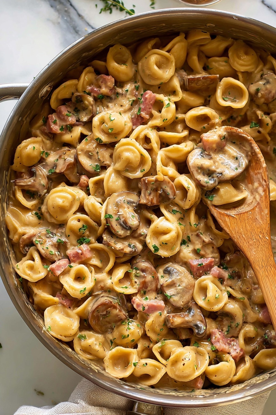 The image shows a close-up view of a pan filled with creamy pasta made from small round orecchiette noodles mixed with sliced brown mushrooms and small pieces of pink ham. The sauce is light brown and smooth, coating every piece of pasta and mushroom evenly. The dish is garnished with small green herb bits scattered on top. A wooden spoon is resting inside the pan, partially covered by the creamy pasta. The pan sits on a white marbled surface with a light-colored cloth nearby. Photo taken with an iphone --ar 2:3 --v 7