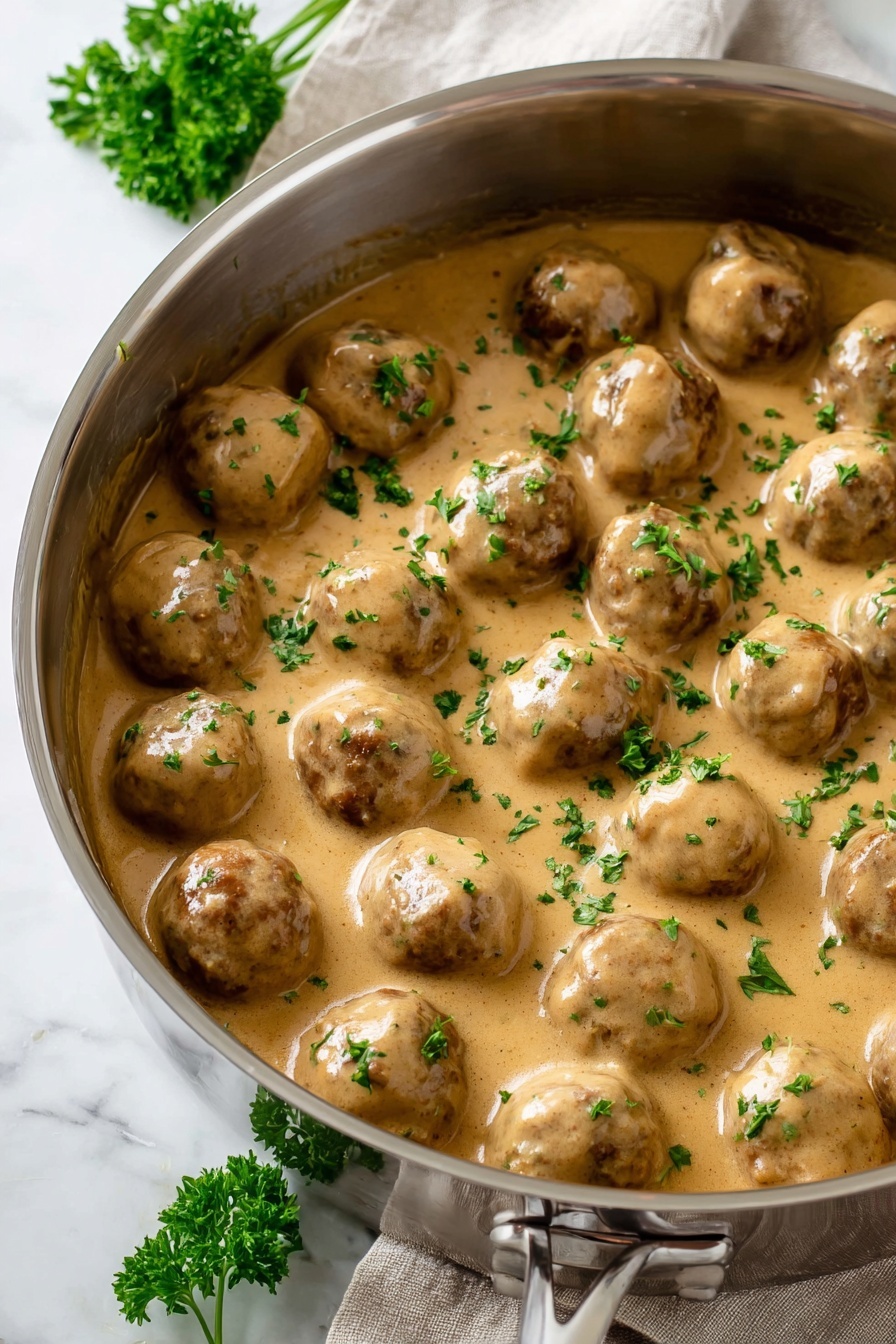 A stainless steel pot filled with around twenty round meatballs covered in thick light brown creamy sauce with a smooth texture; small bright green parsley leaves are sprinkled evenly over the top, adding color contrast; the pot sits on a white marbled surface with a sprig of parsley on the side and a beige cloth in the background, bright natural lighting enhances the glossy look of the sauce photo taken with an iphone --ar 2:3 --v 7