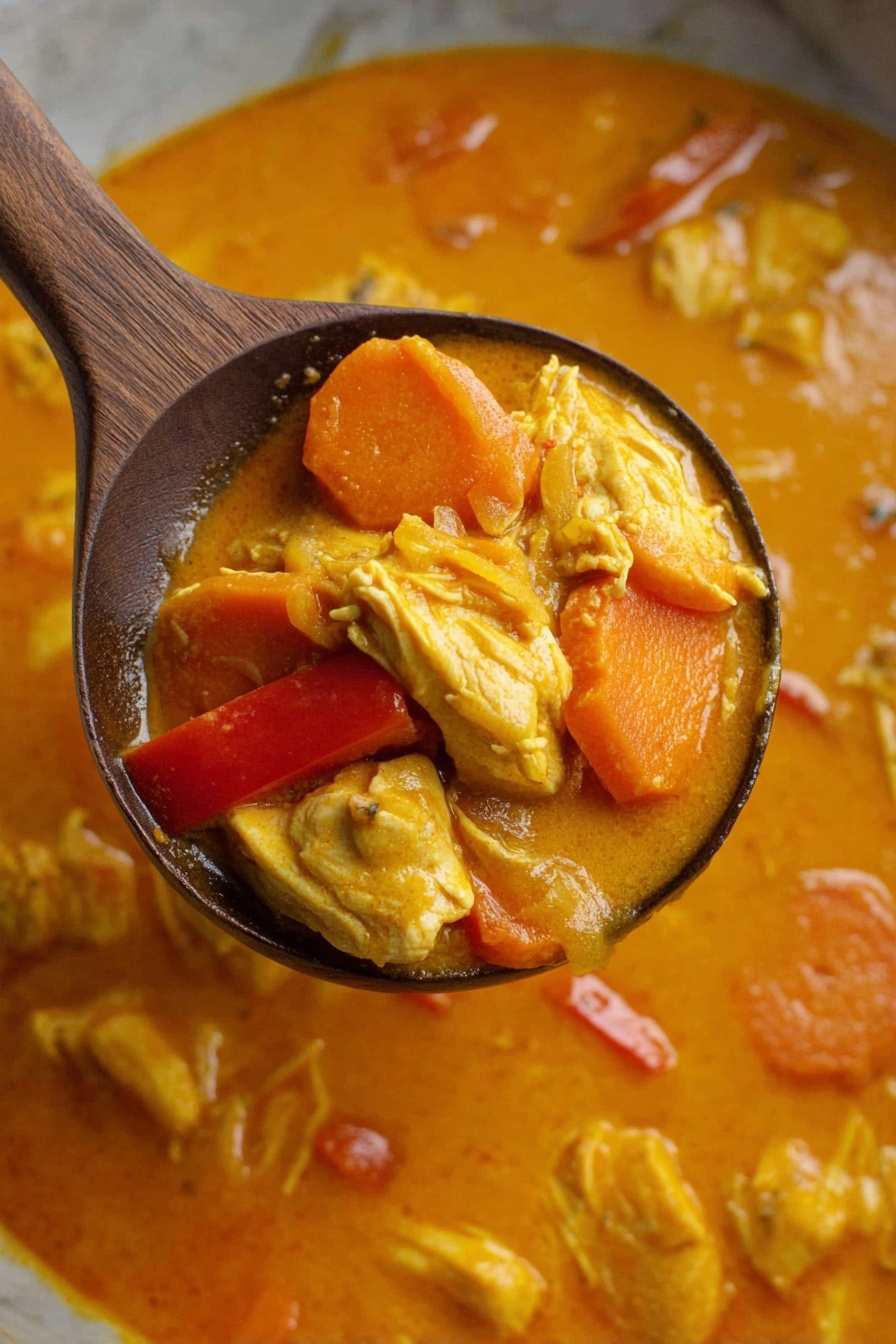 A wooden spoon holds a scoop of thick orange curry with visible layers of yellow chicken pieces, sliced orange carrots, and red bell peppers, all sitting in a creamy orange sauce. The solid ingredients have a soft texture and are mixed together, floating in the sauce that has a smooth and slightly shiny surface. The background shows more of the same curry with small chunks scattered in it, all on a white marbled texture. Photo taken with an iphone --ar 2:3 --v 7