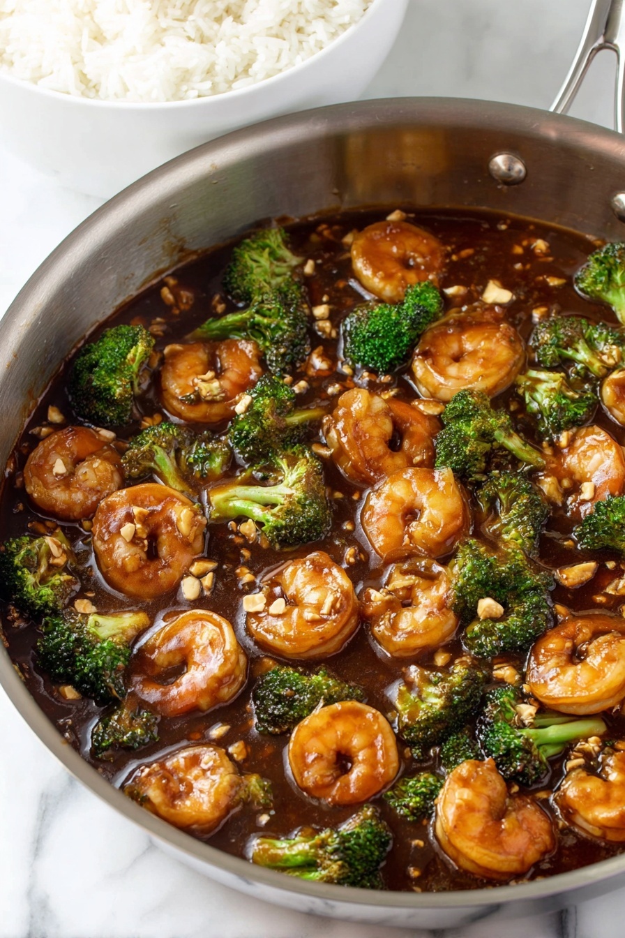 The image shows a stainless steel pan filled with shrimp and broccoli pieces cooked in a thick, dark brown sauce. The shrimp are plump and glazed, spread evenly throughout the pan, with small broccoli florets scattered between them. The sauce has a shiny appearance and small bits of what looks like chopped garlic or nuts sprinkled on top. In the background, there's a partial view of a white bowl filled with plain white rice. The whole setup is placed on a white marbled surface photo taken with an iphone --ar 2:3 --v 7