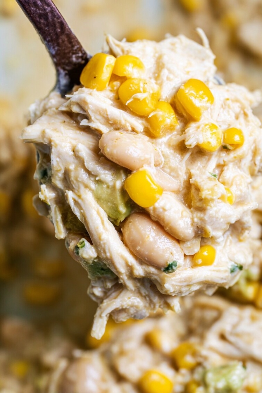 A close-up of a creamy mixture with a thick texture, showing shredded white chicken pieces and plump light beige beans mixed throughout. Bright yellow corn kernels are scattered on top, adding a pop of color against the creamy, slightly off-white base. The mixture looks smooth and rich, with the ingredients evenly blended. A dark spoon scoops a portion of this dish with a blurred background that hints at more of the same mixture. The photo was taken on a white marbled surface. photo taken with an iphone --ar 2:3 --v 7
